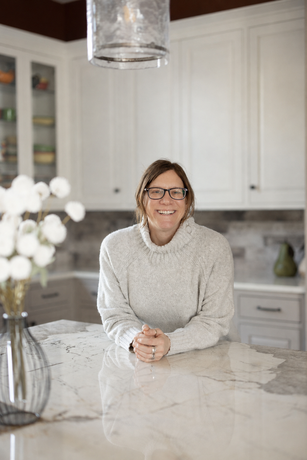 Woman smiling and leaning on a kitchen counter, wearing glasses and a white sweater.