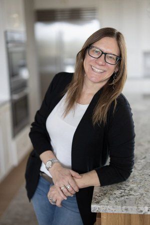 Woman smiling and leaning on a kitchen counter, wearing glasses, black cardigan, and white top.