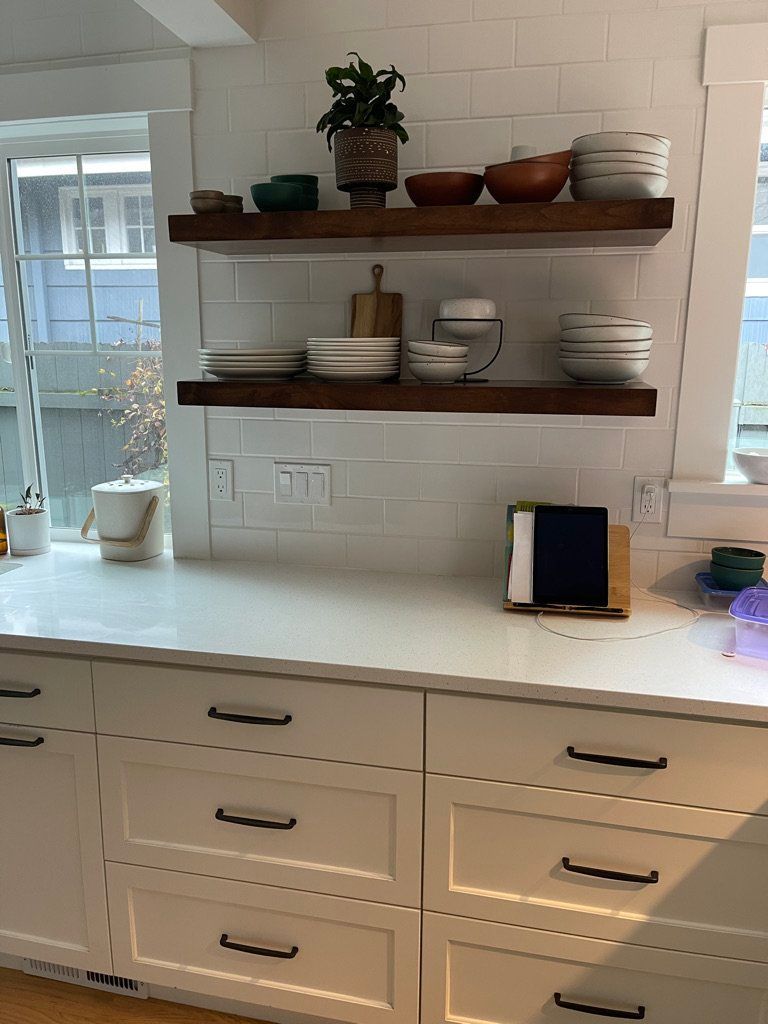 Kitchen counter with white cabinets, white tile backsplash, and wooden shelves holding dishes and plants