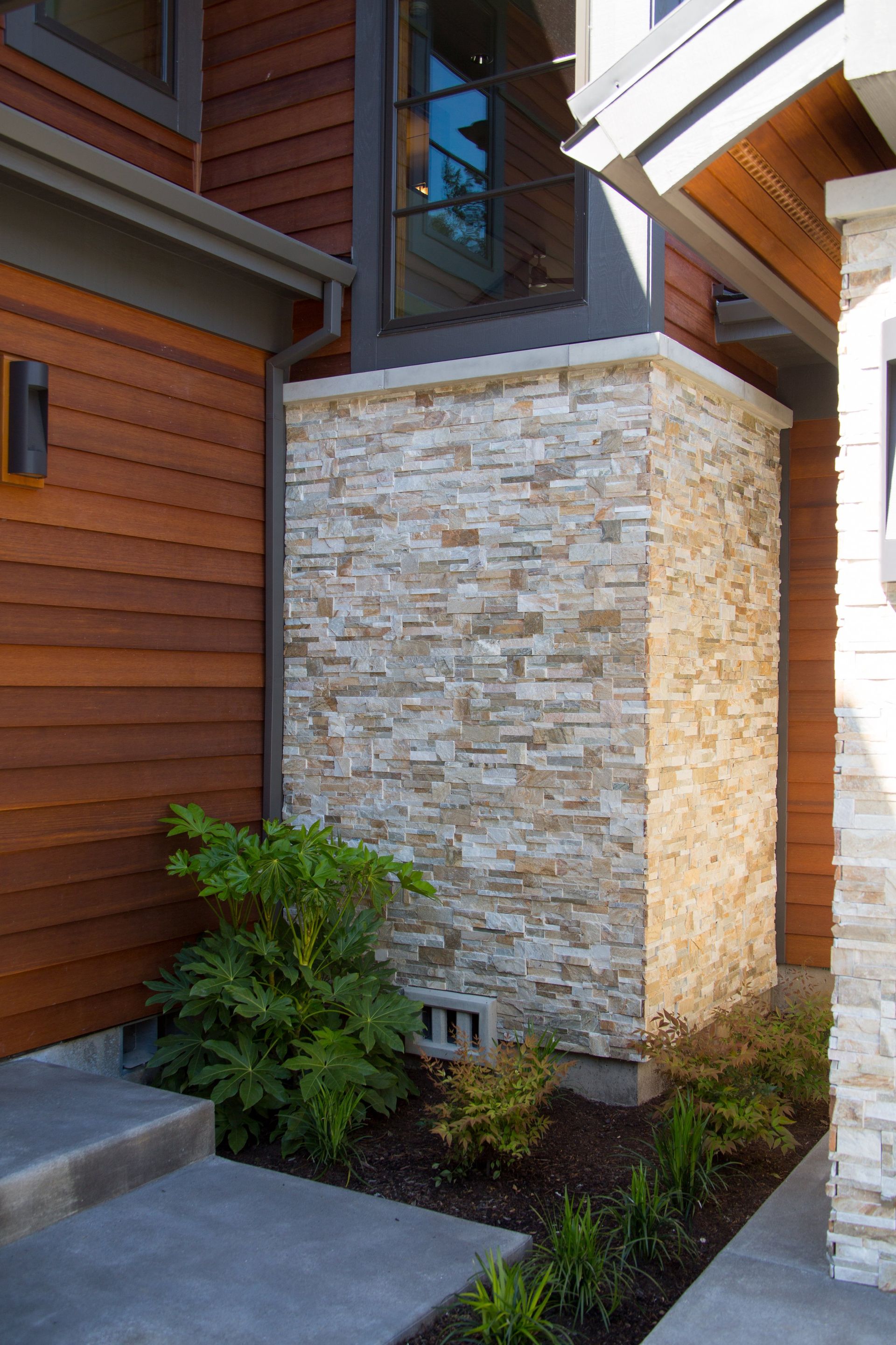 Landscaped corner beside a house with stacked stone column, brown siding, and small shrubs.