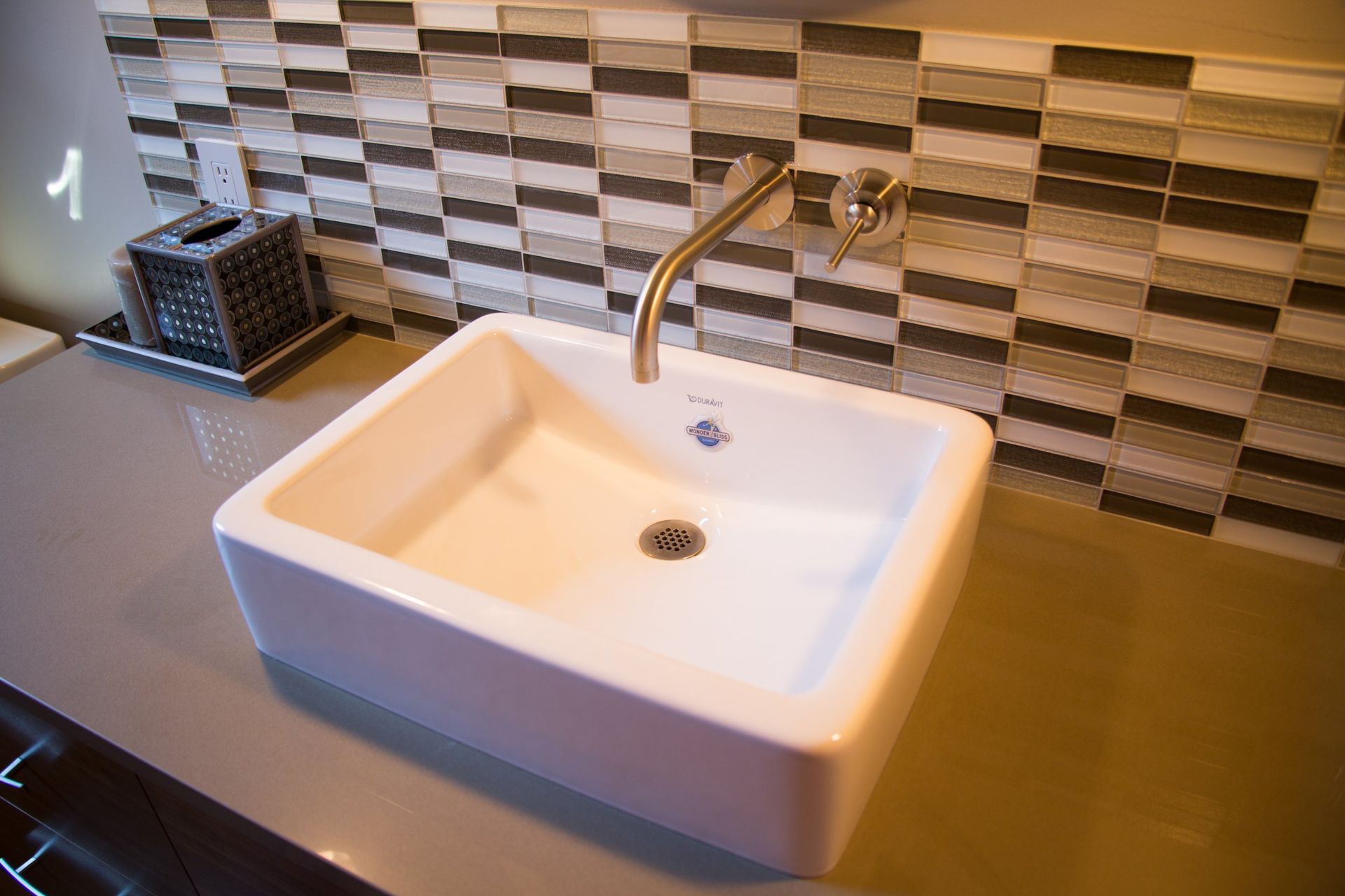 White square sink with chrome faucet against a beige tiled backsplash on a dark countertop.
