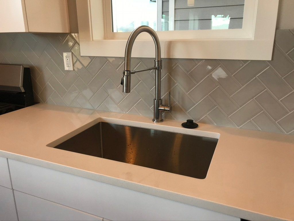 Modern kitchen sink with stainless basin, tall faucet, beige countertop, and gray tile backsplash