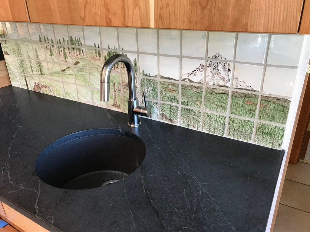 Kitchen sink with chrome faucet, black countertop, and tiled backsplash with landscape design