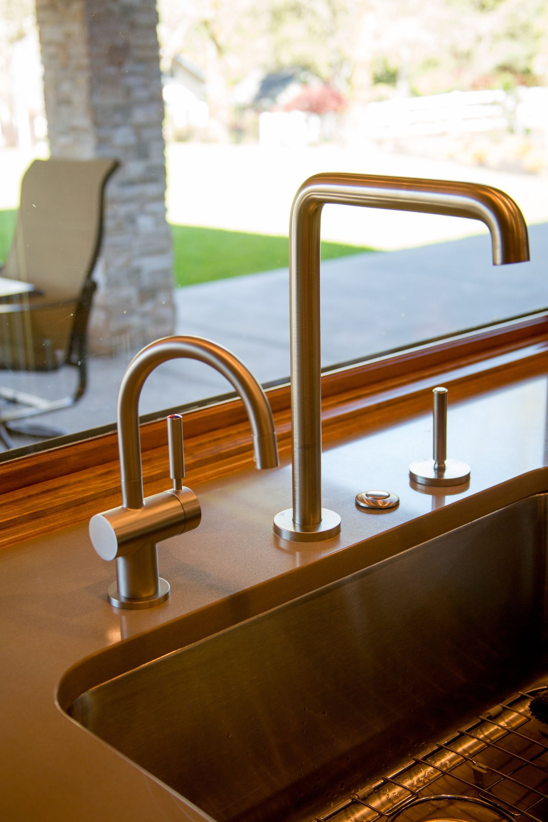 Bronze kitchen faucet and soap dispenser beside a sink, with a bright patio visible beyond the window