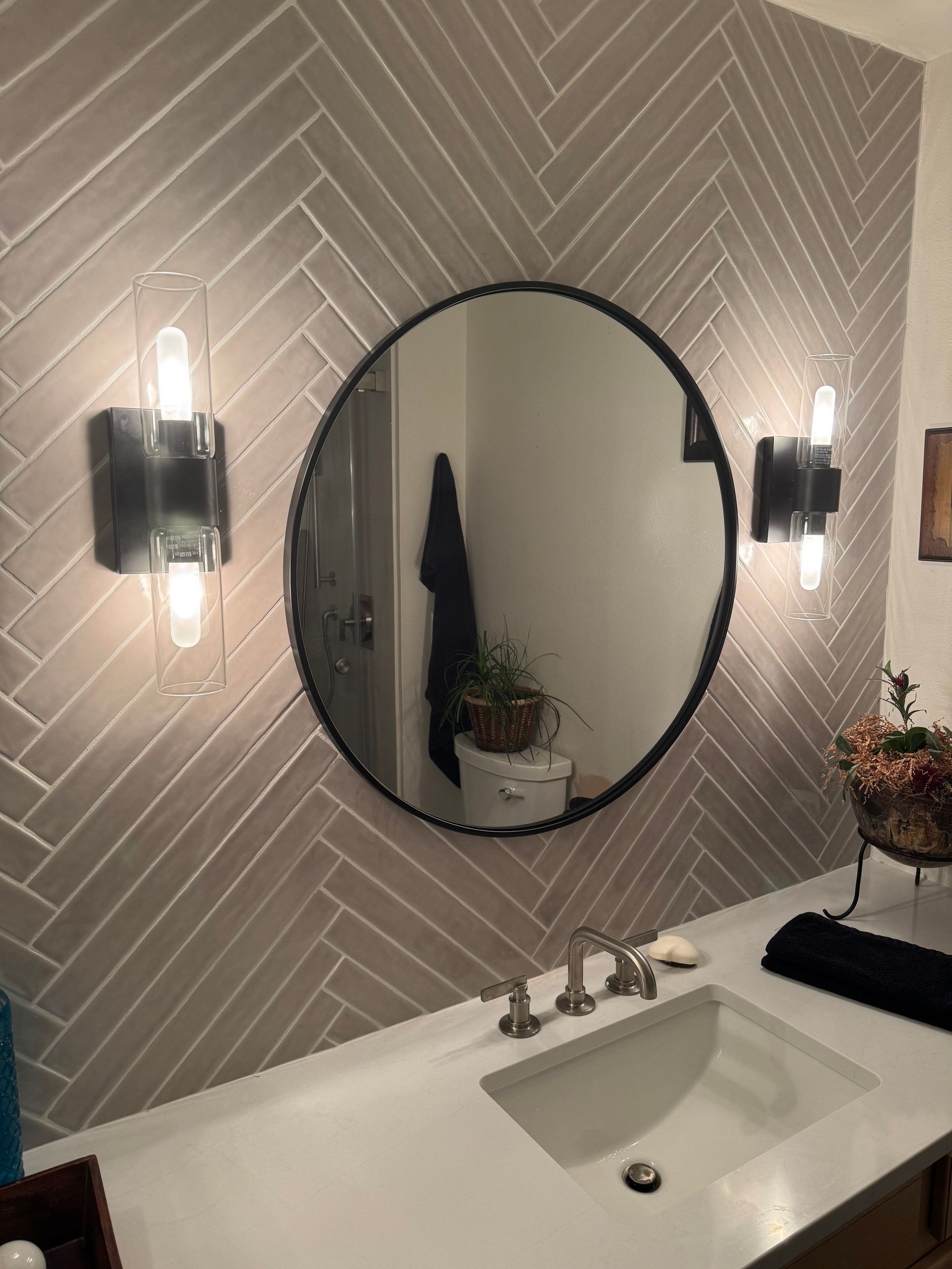 Bathroom vanity with round mirror, wall sconces, and white sink on a herringbone tile wall.