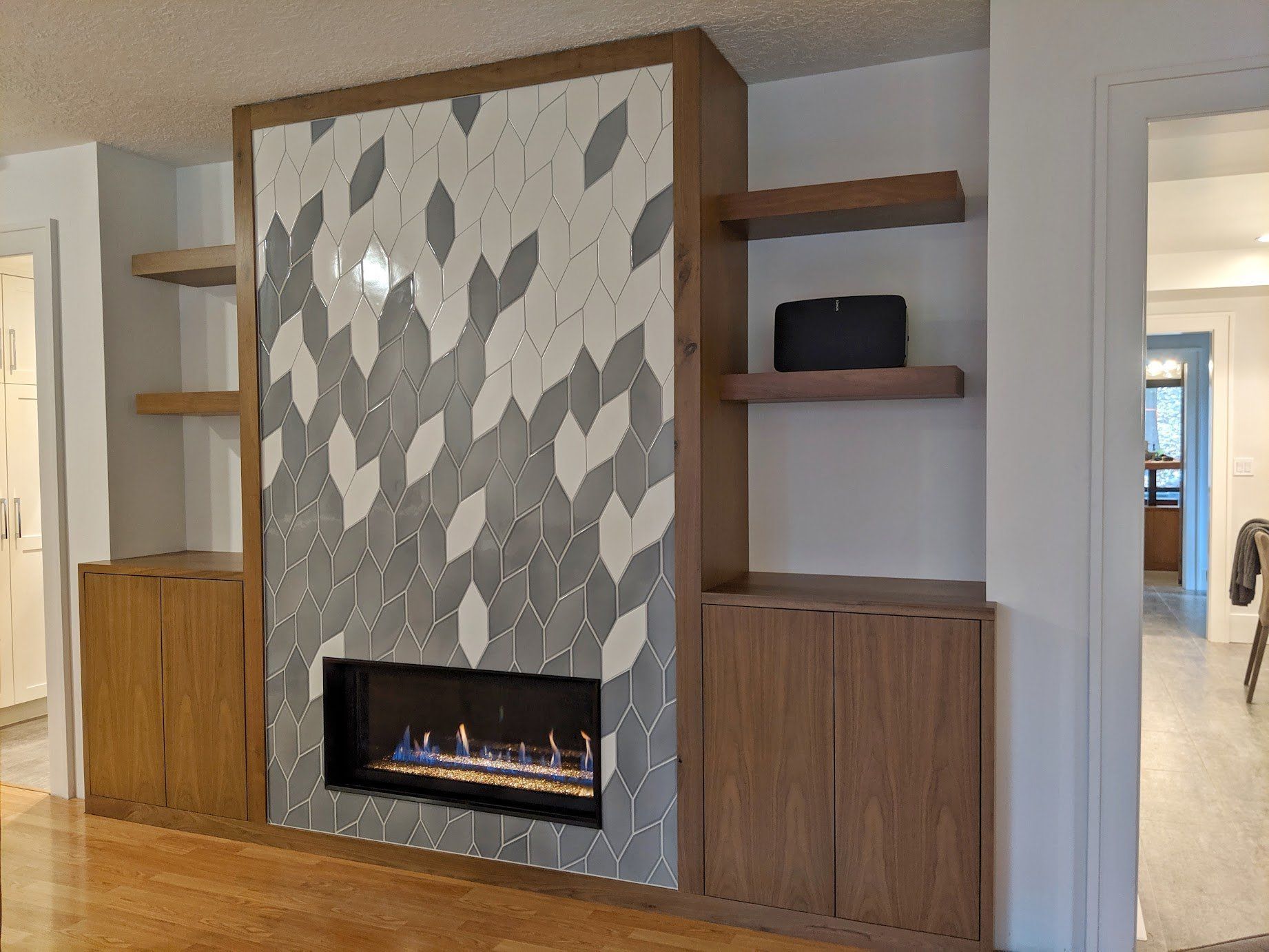 Modern living room fireplace with patterned tile surround, wood shelving, and wooden cabinetry.