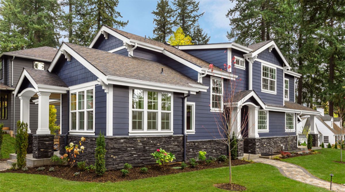 Gray-blue suburban house with white trim, stone accents, and manicured lawn under tall trees