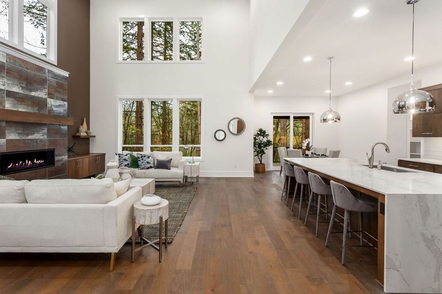Bright open-concept living room with white sofas, wood floors, tall windows, and a kitchen island with bar stools