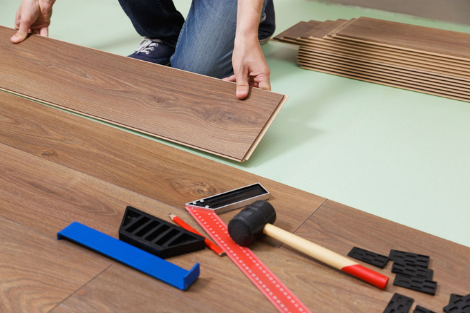 Hands installing a laminate floor plank with tools and spacers on the floor