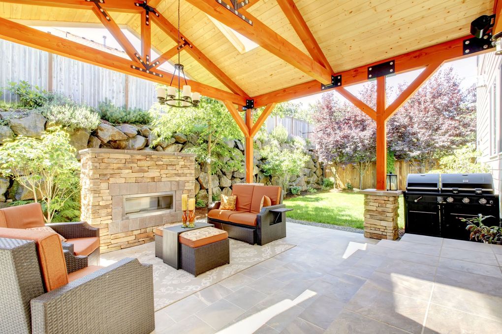 Covered patio with stone fireplace, wicker seating, and a black grill in a landscaped backyard