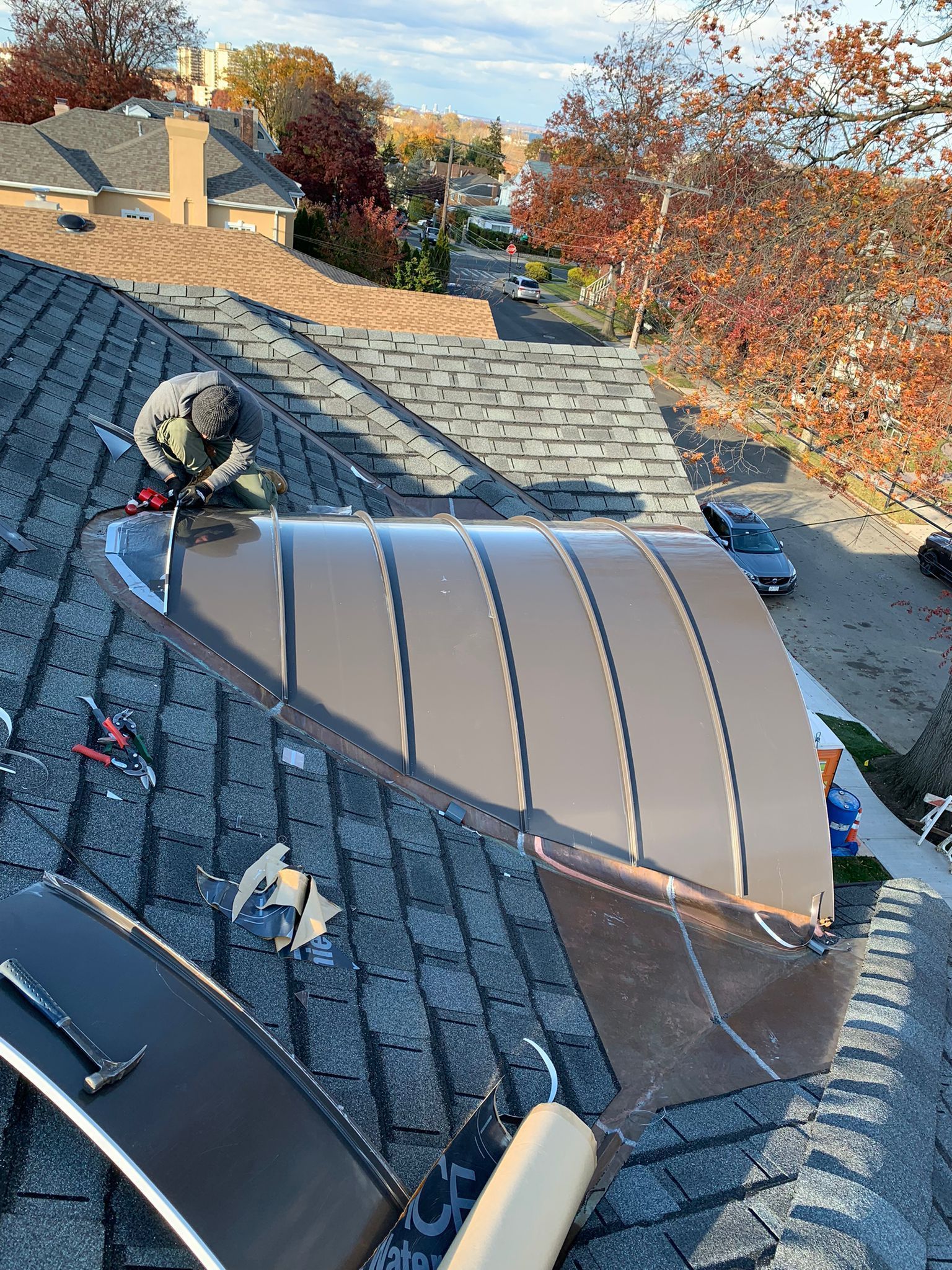 A person installs a curved, brown metal vent cover onto a shingled roof on a sunny day.