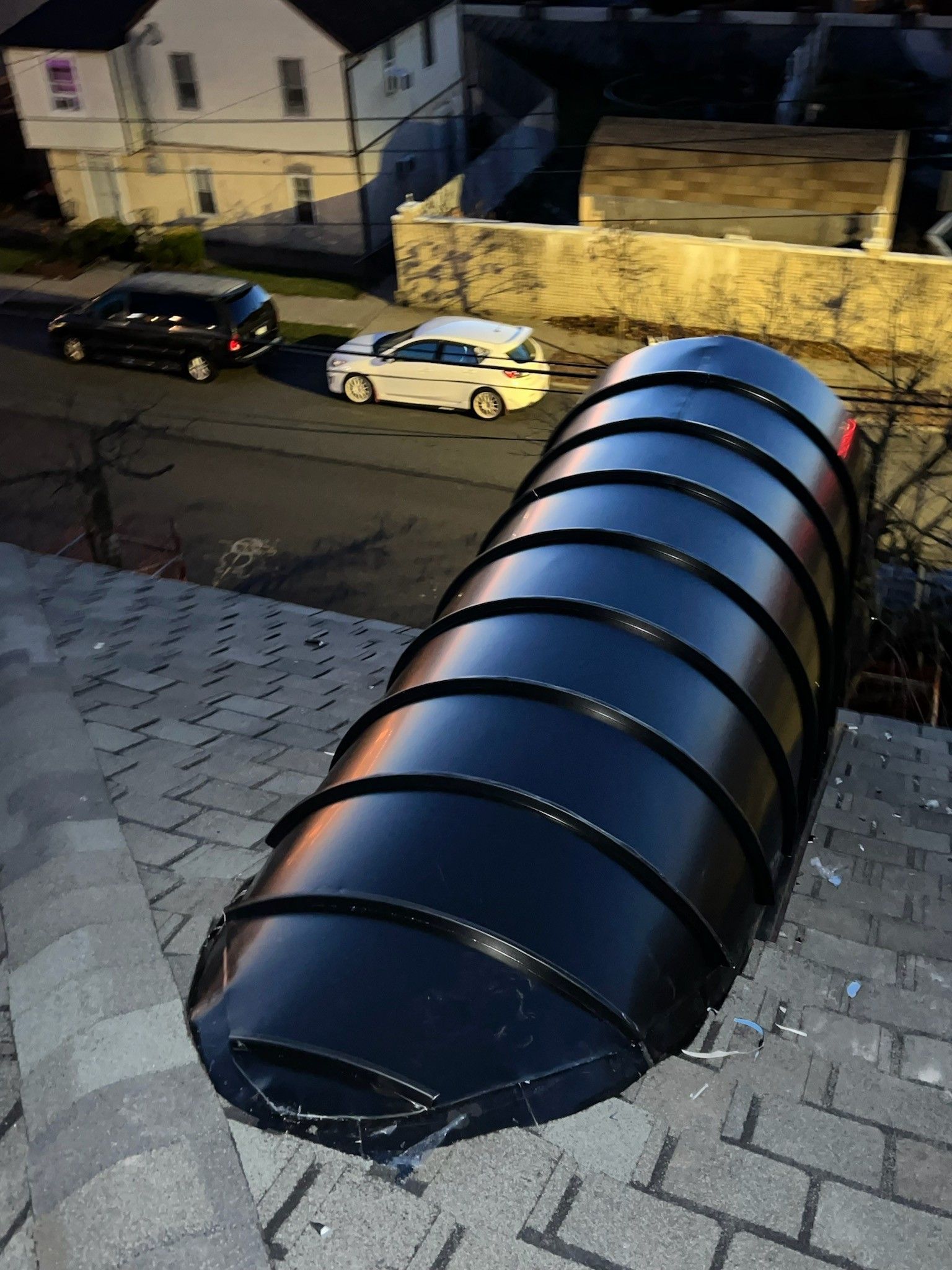 A large, black, corrugated metal tunnel structure mounted on a shingled roof, with cars and a house visible below.
