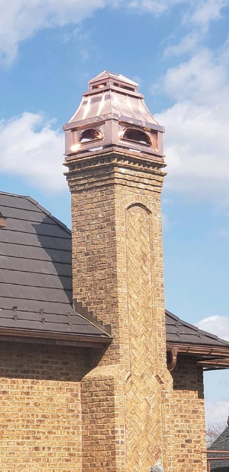 An ornate brick chimney with a decorative metal-covered top, rising above a shingled roof against a blue sky.