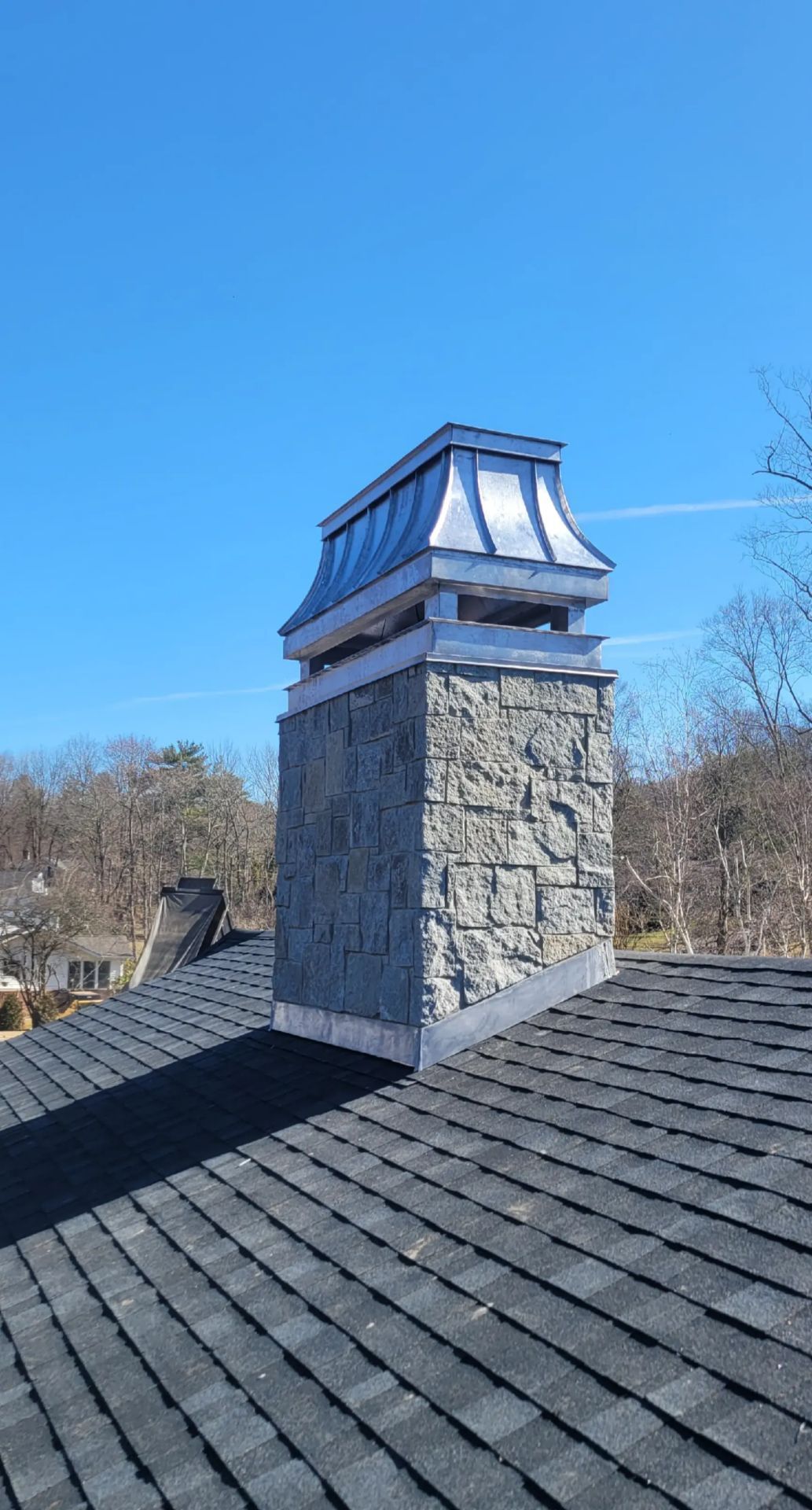 A stone chimney with a metal cap stands on a shingled roof under a clear blue sky.
