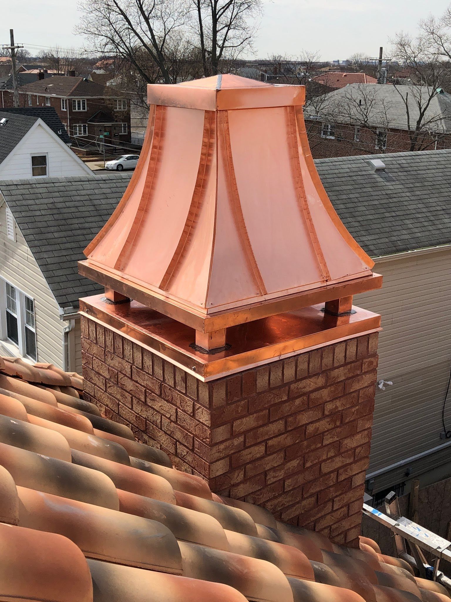 A copper chimney cap with a flared, pagoda-style top sits on a brick chimney on a roof with terracotta-colored tiles.