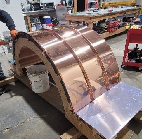 A person installs copper sheeting onto a curved wooden frame in a workshop.