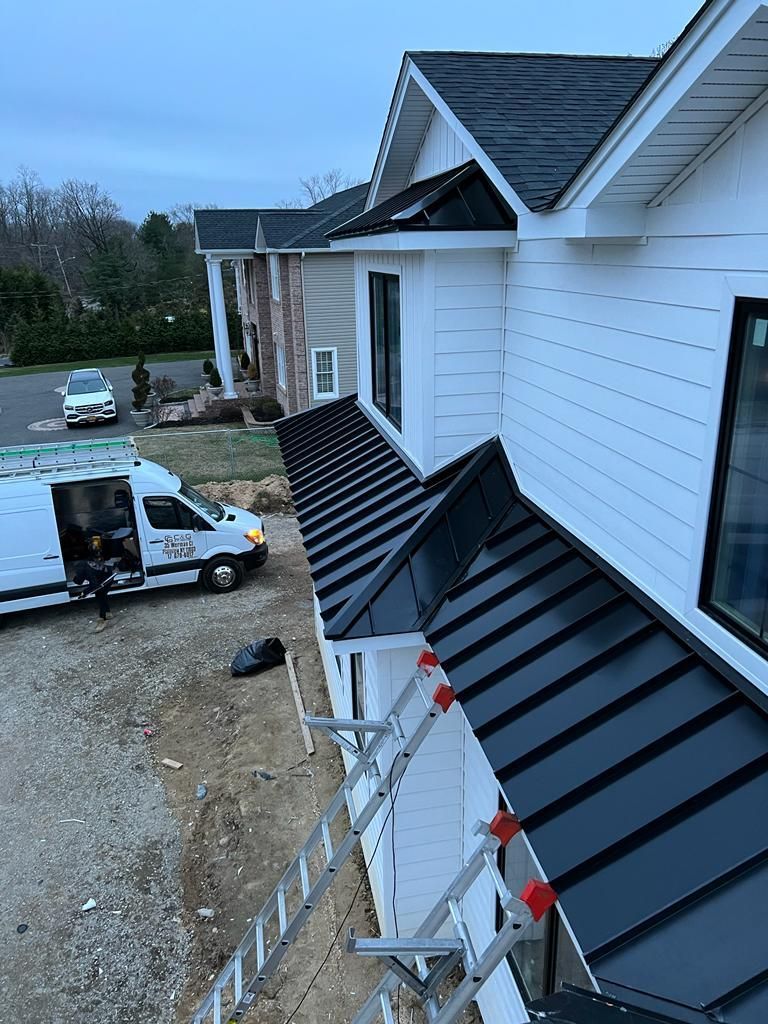 A white house under construction with a new black standing seam metal roof and siding, next to a work van and ladder.