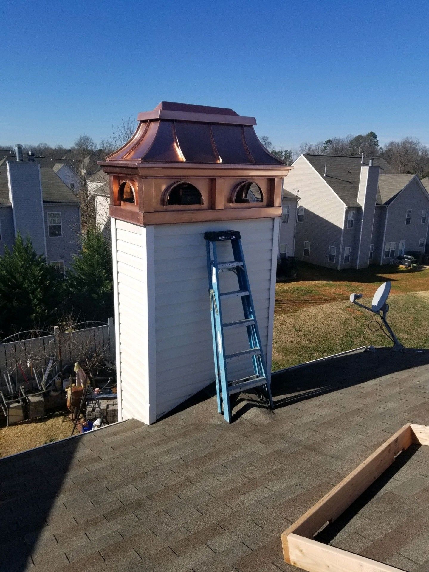 A copper-topped chimney shroud sits on a residential rooftop with a blue ladder leaning against the white-sided chase.