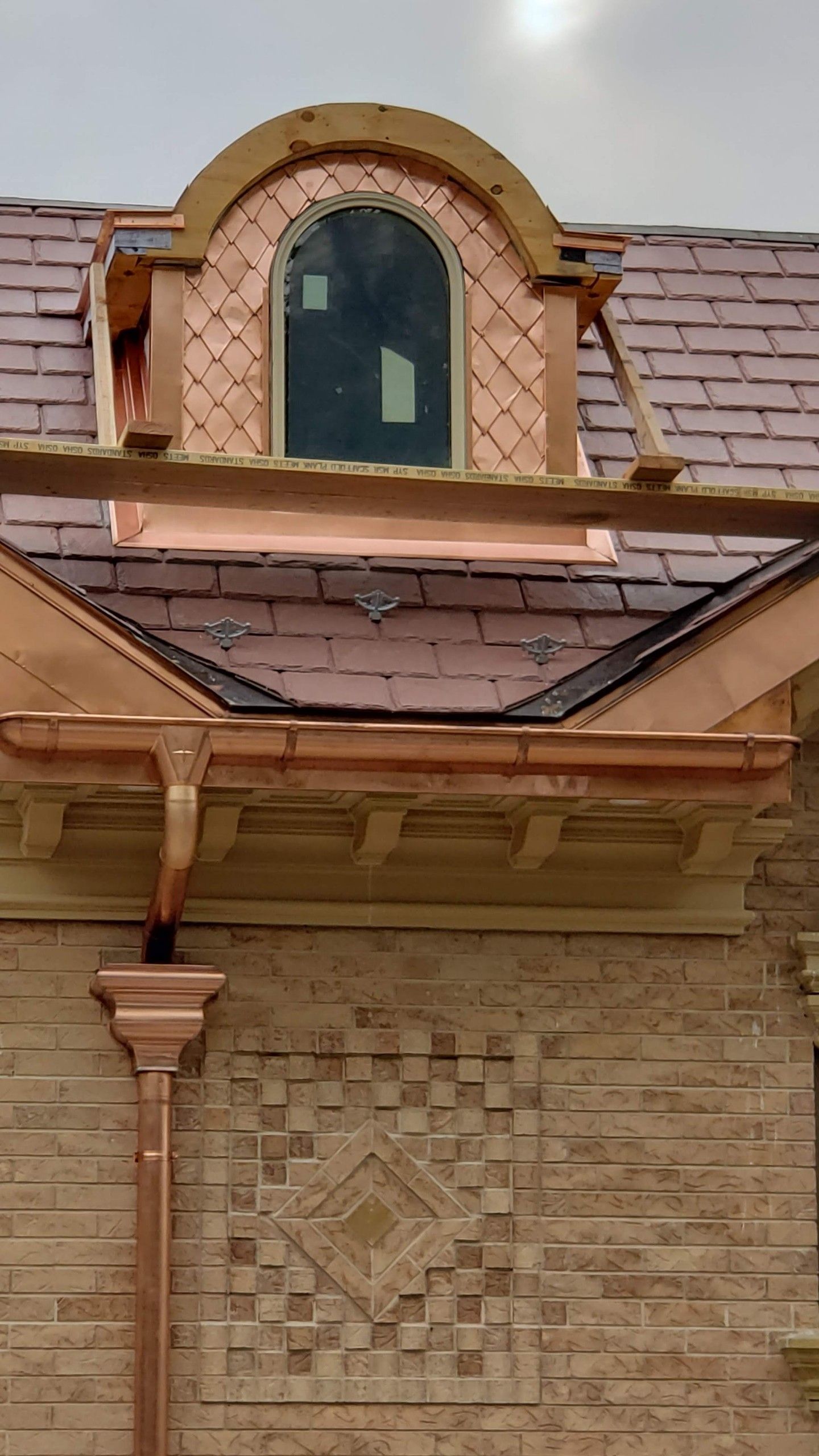 A copper-clad arched dormer window set into a slate tile roof above a brick wall with a decorative inset diamond pattern.