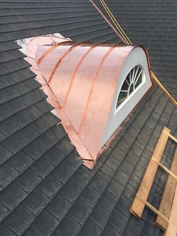 A curved, copper-clad dormer window set into a dark gray shingled roof under construction.