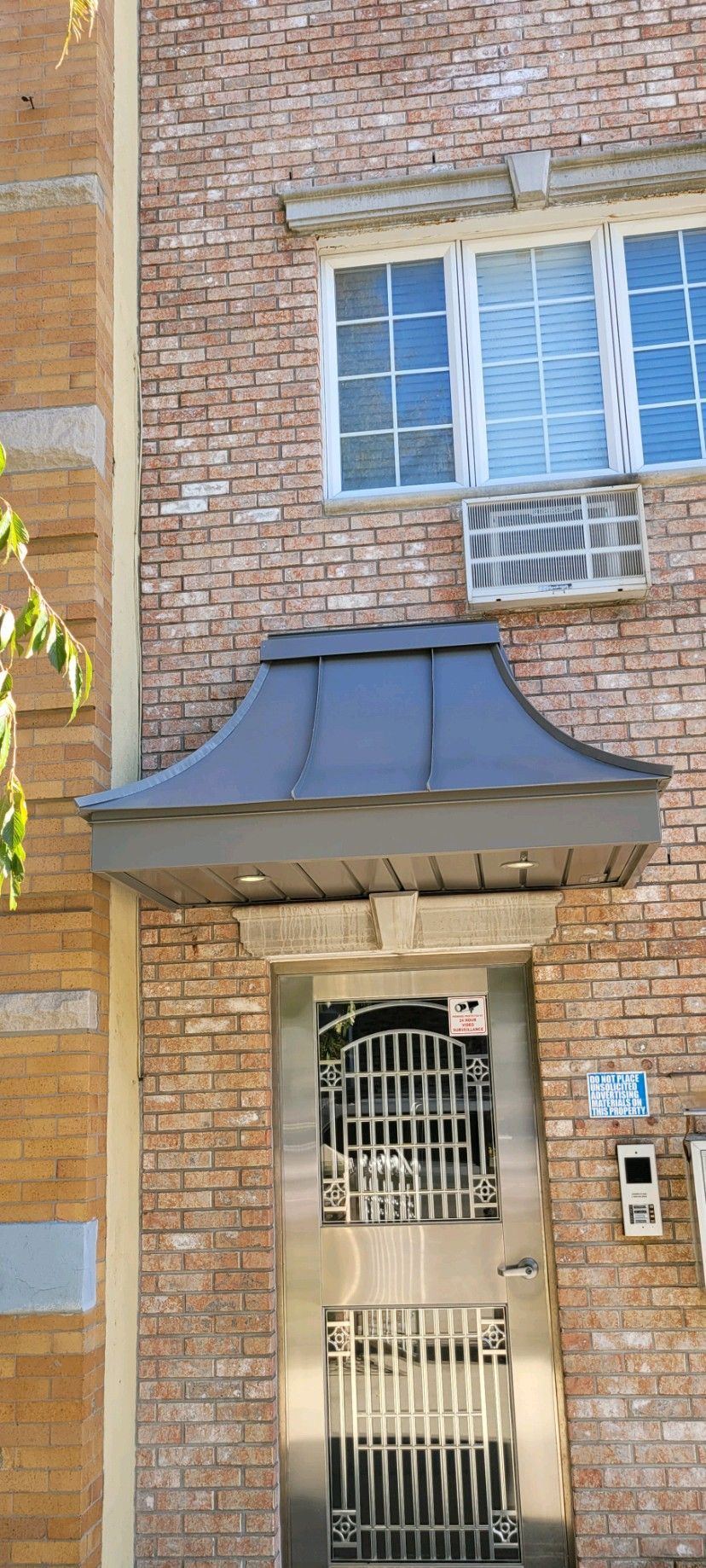 A brick building entrance featuring a dark metal curved awning over a secure, decorative stainless steel door.
