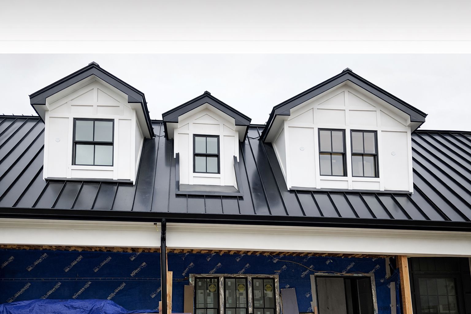 A house exterior under construction, featuring a black metal roof with three white dormer windows and blue wall sheathing.