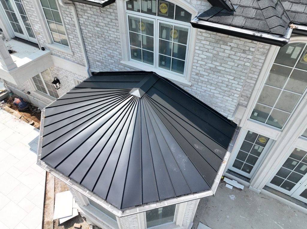 An aerial view of a dark metal, multi-faceted roof covering a bay window section on a light-colored brick home.