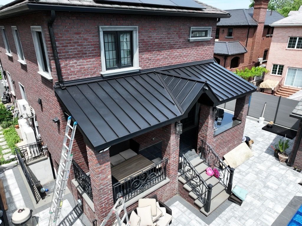 A two-story brick house with a newly installed black metal roof over an entryway and patio, captured from a high angle.