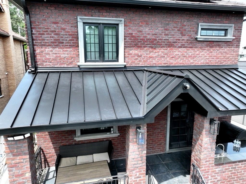 A two-story brick house with a dark metal roof covering a front porch, featuring a bench and stone columns.