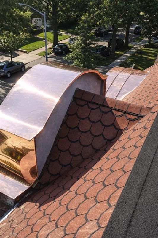 An overhead view of a copper-clad arched dormer roof transition next to red scalloped shingles on a sloped roof.