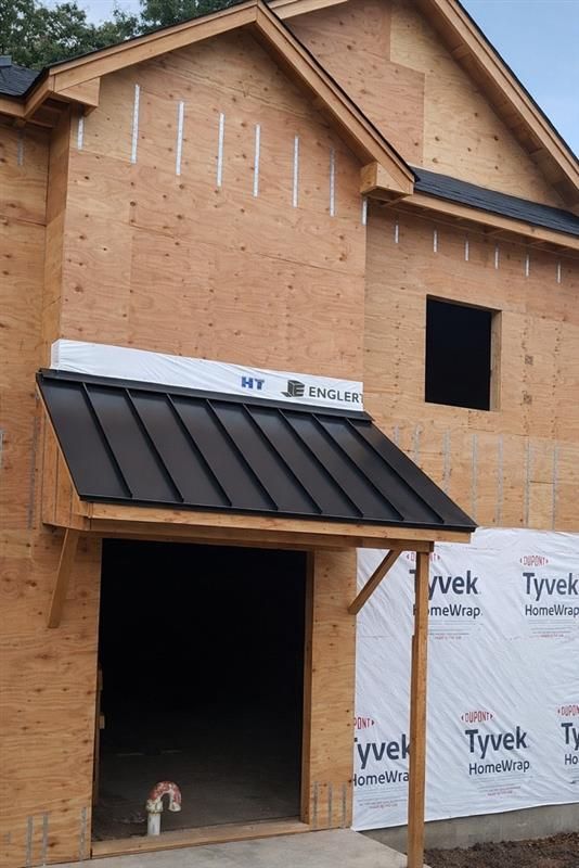 Construction site showing a house frame with plywood walls, Tyvek house wrap, and a black metal awning over the doorway.