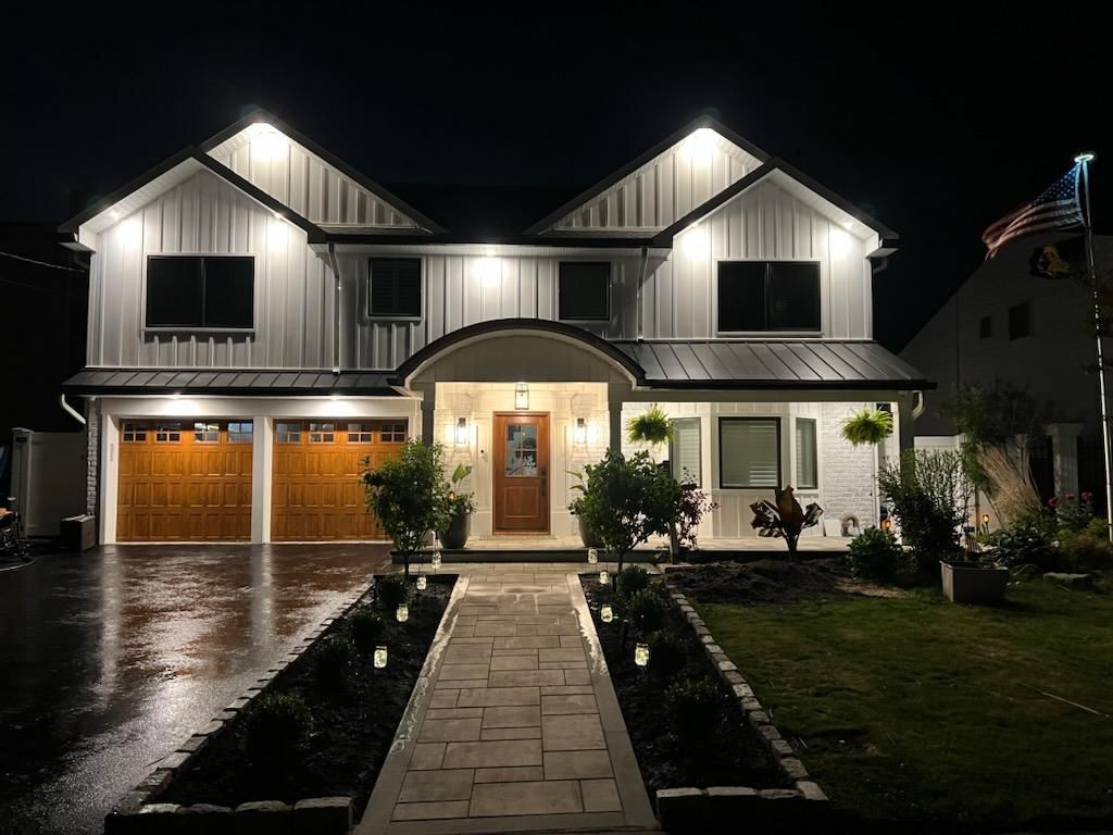 A modern two-story home at night with white vertical siding, two wooden garage doors, and a stone walkway.