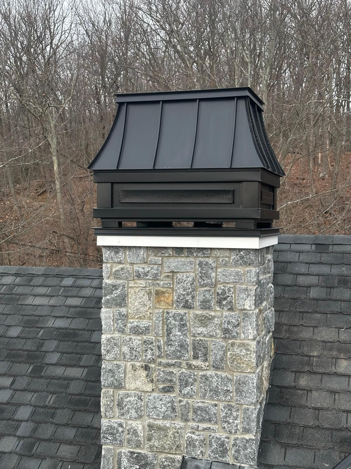 Black chimney cap on a stone chimney. Gray roof, trees in background.