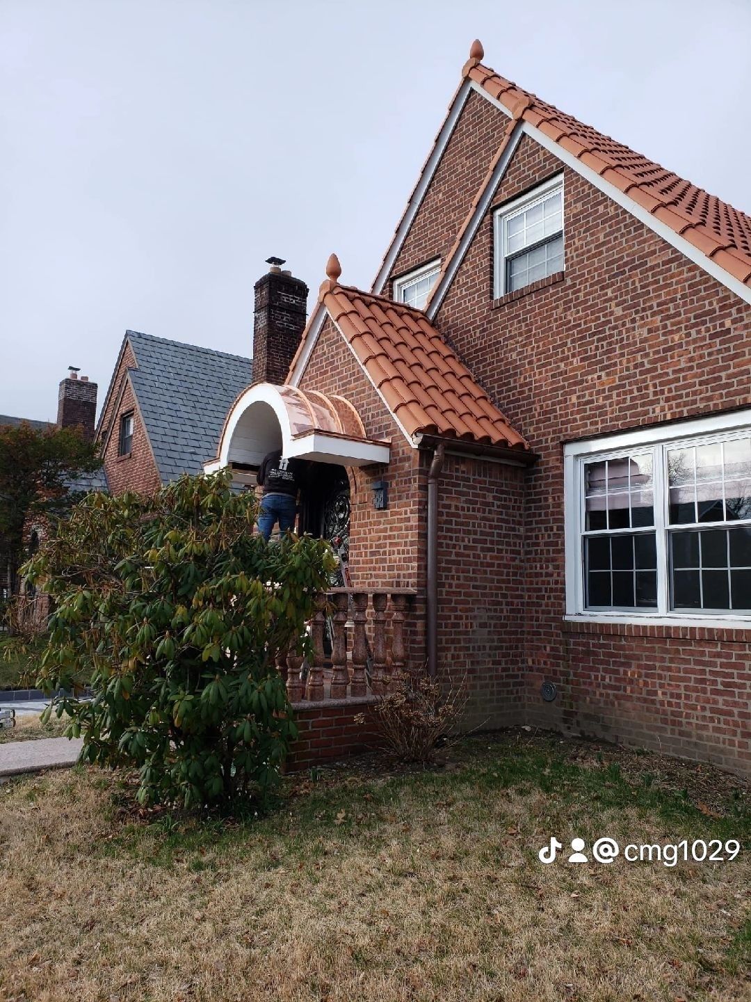 Brick house with red-tiled roof, a person on porch. Overcast sky.