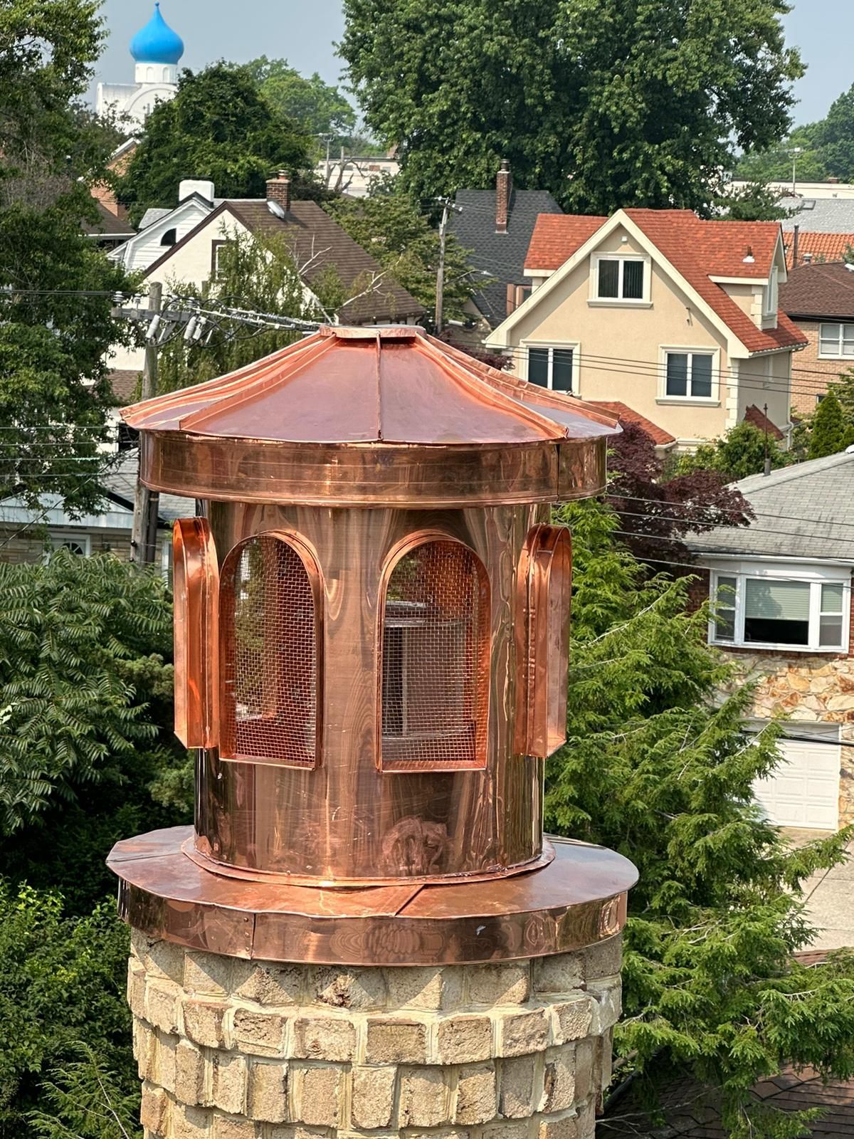 Copper chimney cap with a decorative roof, on a brick chimney, surrounded by green trees and houses.