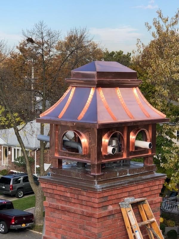Copper chimney cap atop a brick chimney, with arch-shaped vents and a copper roof. Autumn trees in the background.
