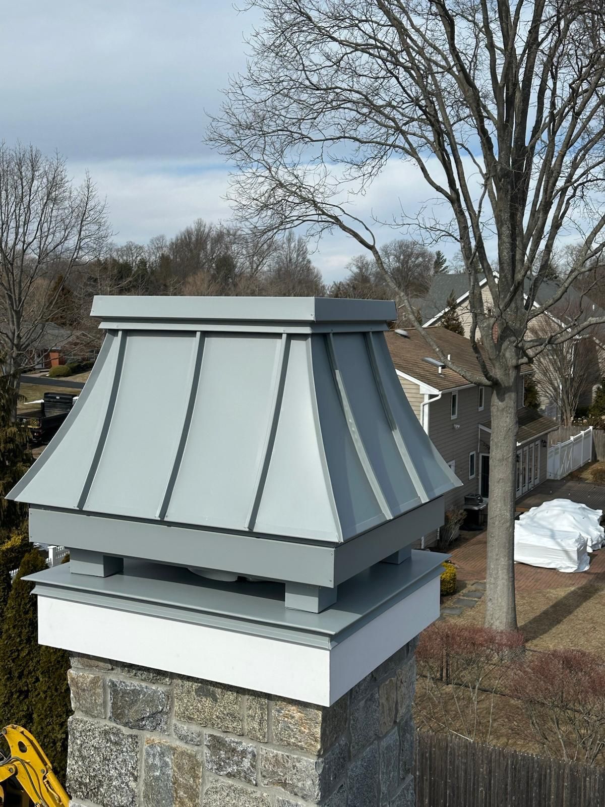 Gray metal chimney cap on a brick chimney, against a cloudy sky and leafless trees.