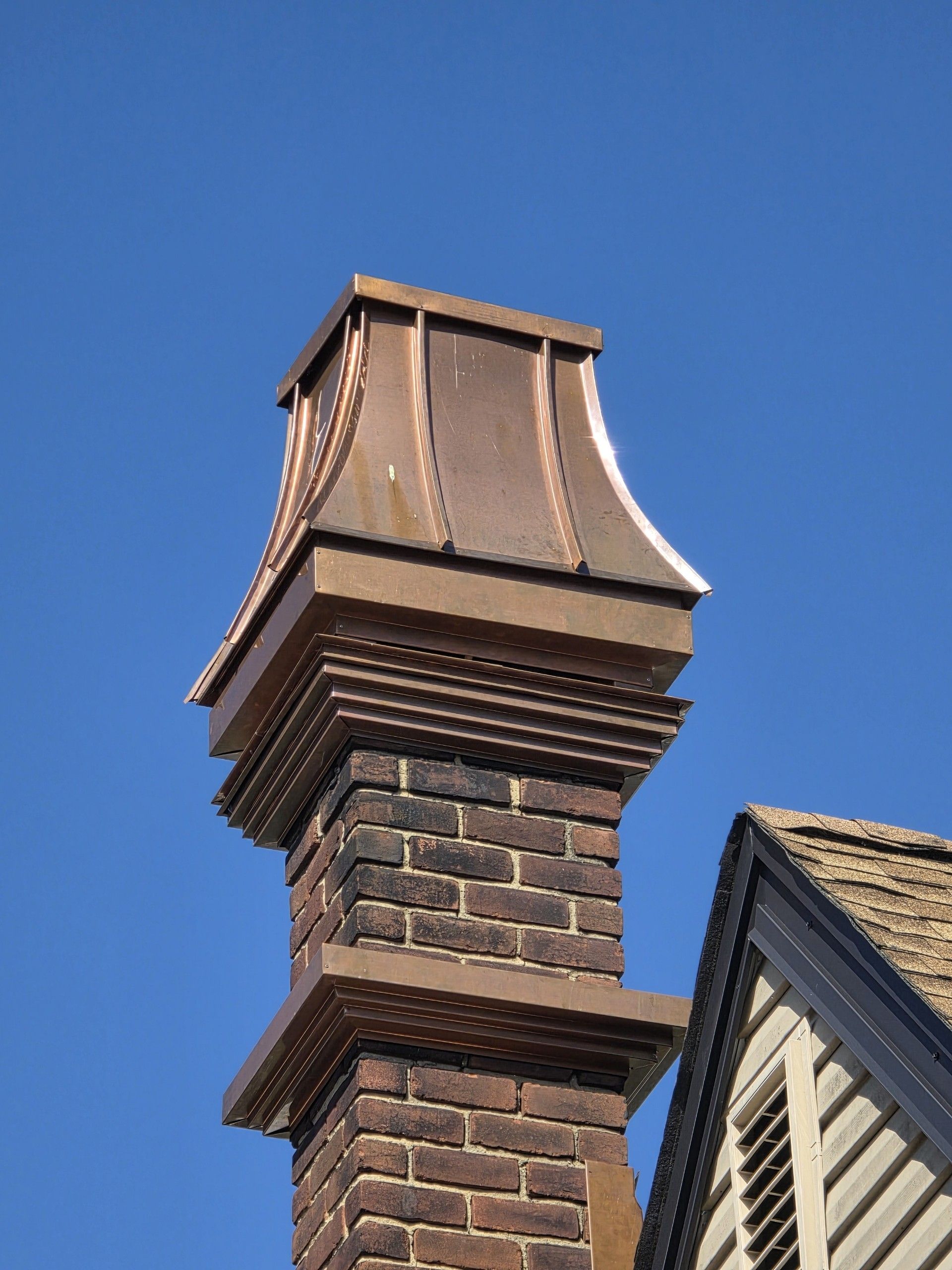 Copper-topped brick chimney against a blue sky, next to a portion of a roof.