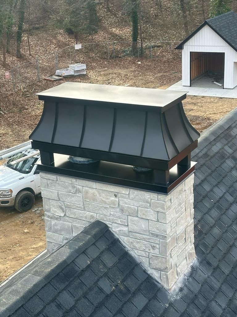 Stone chimney with black metal cap, on a dark shingle roof, with a white truck and a small shed in the background.