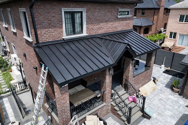 An aerial view of a brick house featuring a newly installed black metal standing-seam roof over the front porch and steps.