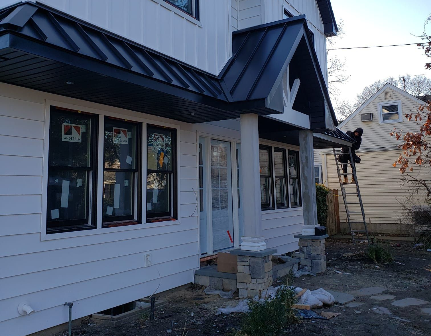 A renovated white house with a dark metal awning over the entrance, where a worker stands on a ladder nearby.