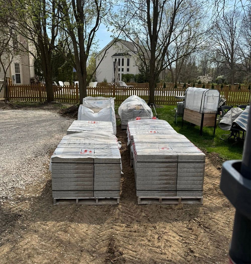 Stacks of wrapped building materials on a gravel lot in front of a house and trees