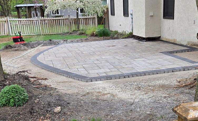 Curved stone patio with dark border beside a house, surrounded by bare dirt and a fenced yard