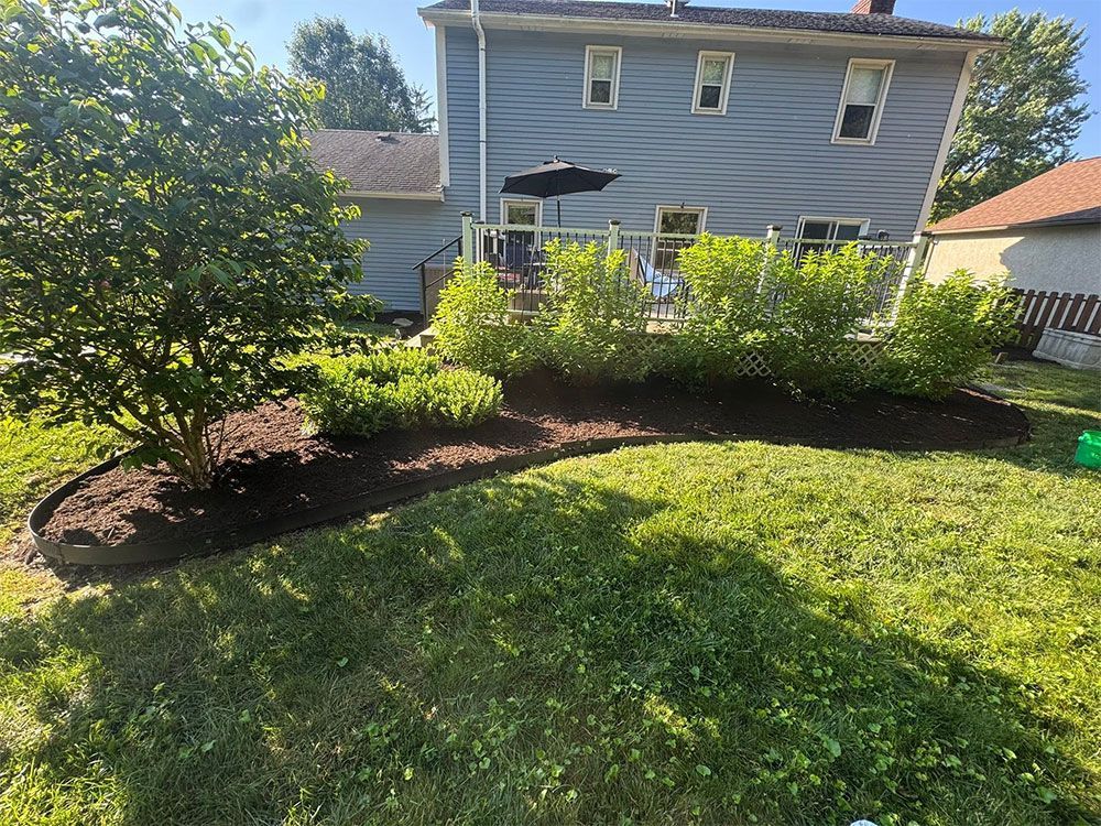 Backyard with green lawn, flower beds, and a gray house with a patio in sunlight