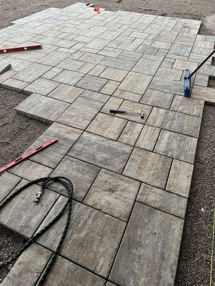 Stone patio under construction with paver tiles, tools, and hoses laid out on a gravel base