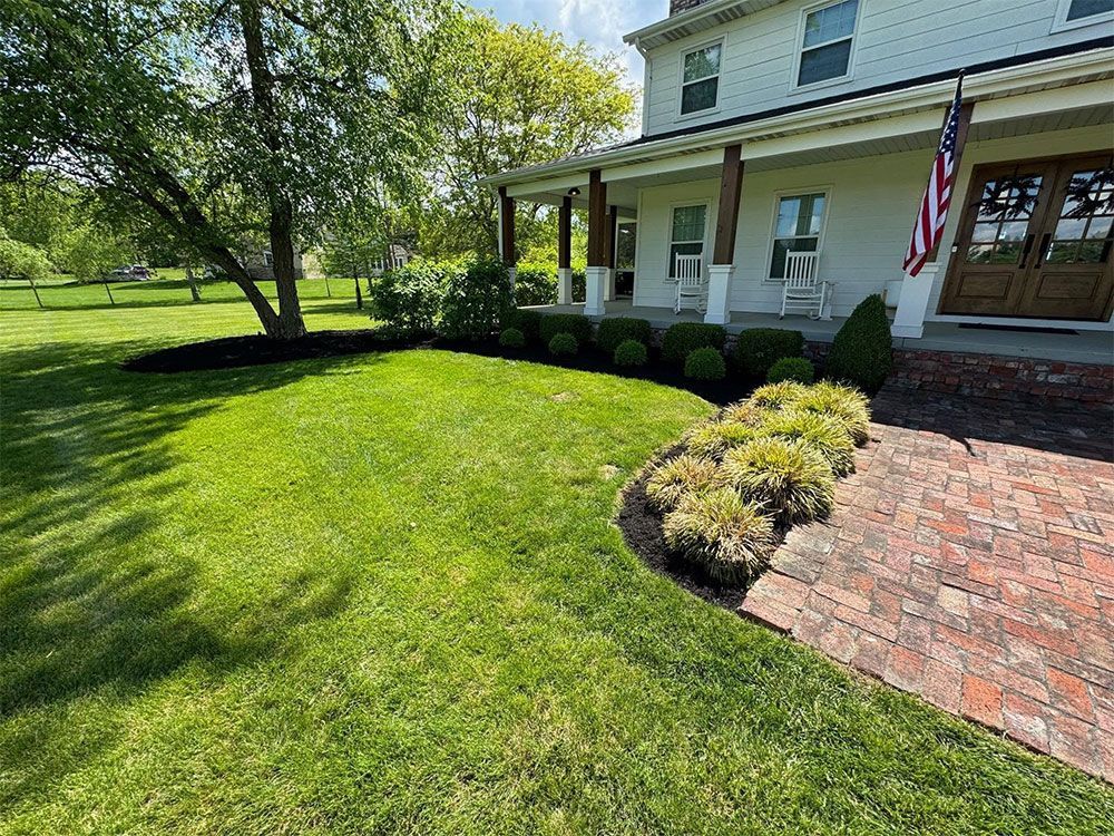 Front yard of a white house with a brick walkway, green lawn, trees, and an American flag on the porch.