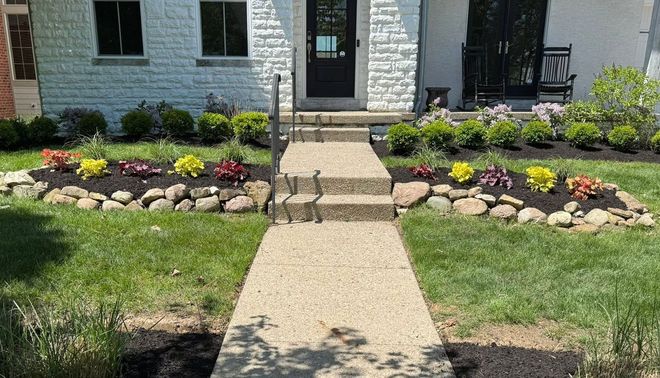 Front walkway to a townhouse with stone steps, black door, and colorful flower beds on both sides.