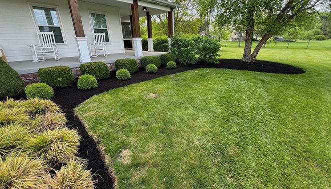 Front yard with a white porch, trimmed shrubs, black mulch border, and a green lawn beside a tree.