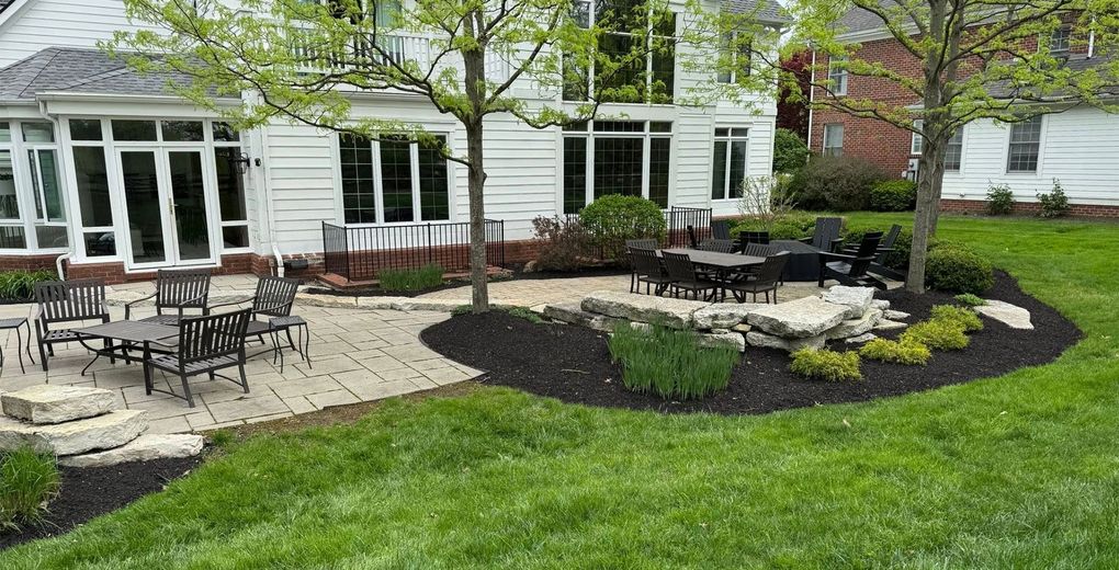 Backyard patio with outdoor tables, stone seating area, and curved mulch bed beside a white house