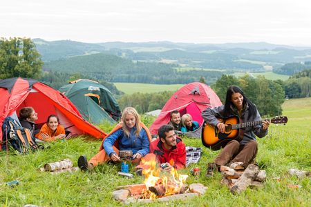 Group of friends camping, gathered around a campfire on a grassy hill, one playing guitar. Tents are in the background with a scenic view.
